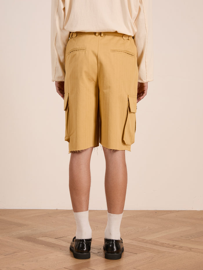 A person stands indoors on a wooden floor, shown from the back, wearing a cream long-sleeve shirt, FOUND Sunlight Twill Cargo Shorts with raw hems in yellow herringbone, white socks, and black shoes.