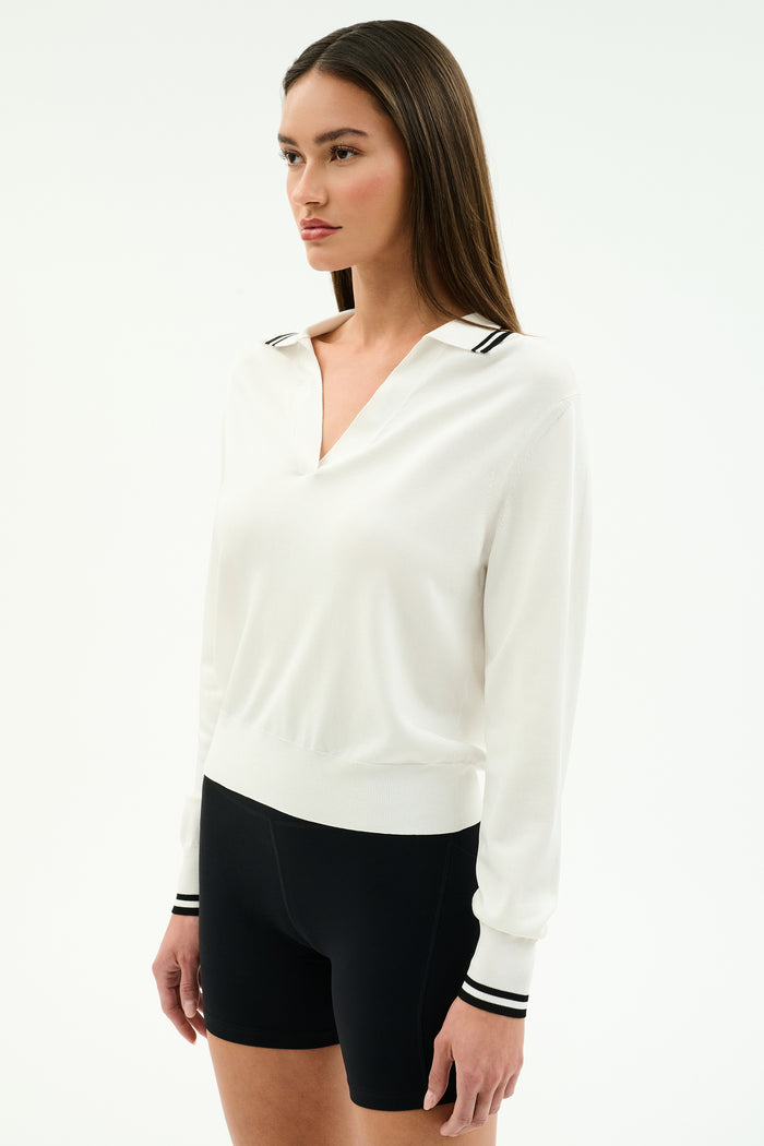 A woman poses against a plain background wearing the SPLITS59 Taylor Knit Polo in white with black stripes, featuring long sleeves and a collared design, styled with fitted black shorts.
