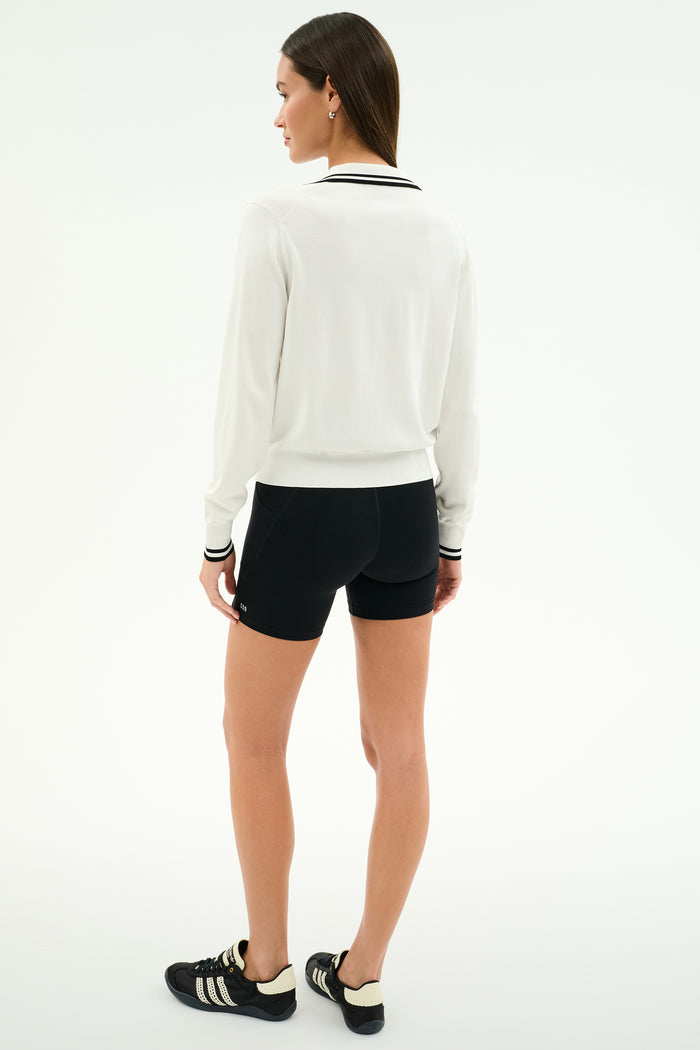A woman faces away, wearing the SPLITS59 Taylor Knit Polo in White/Black, paired with black shorts and black athletic shoes with white stripes, set against a plain light background.