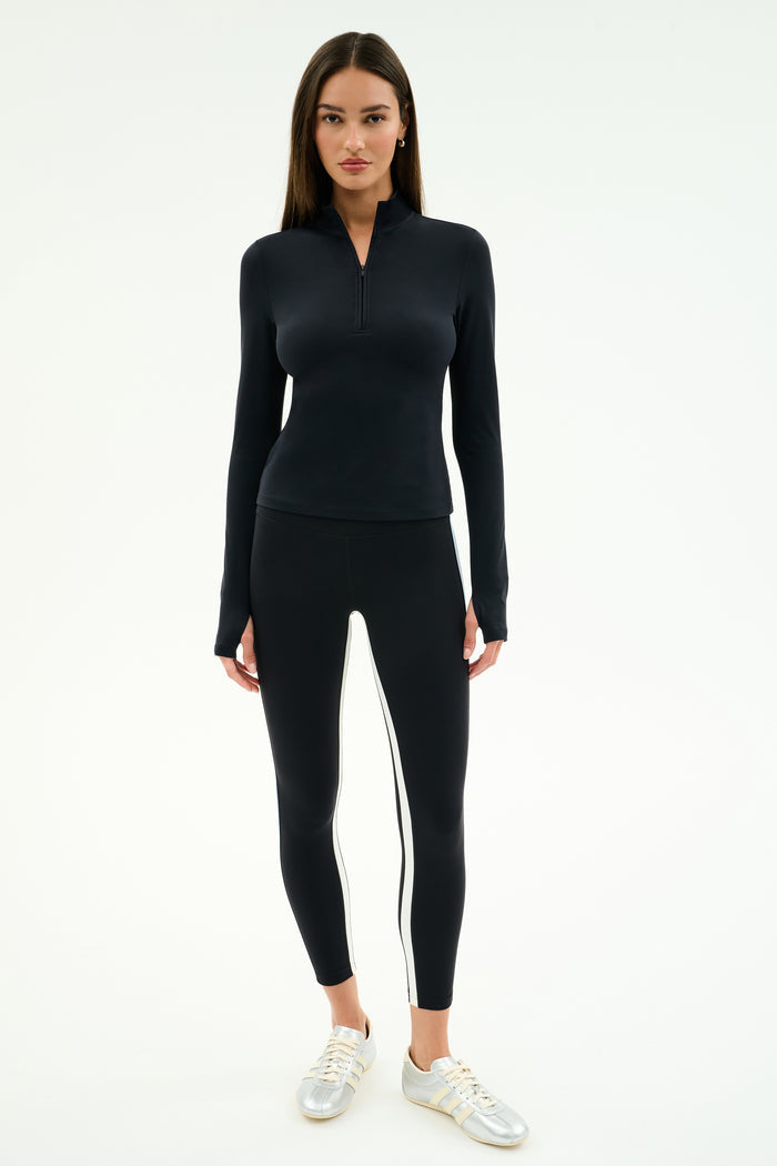 A woman stands facing forward, wearing the SPLITS59 Tess Rigor Half Zip Waist Length top in black, black leggings with white side stripes, and silver sneakers against a plain white background.