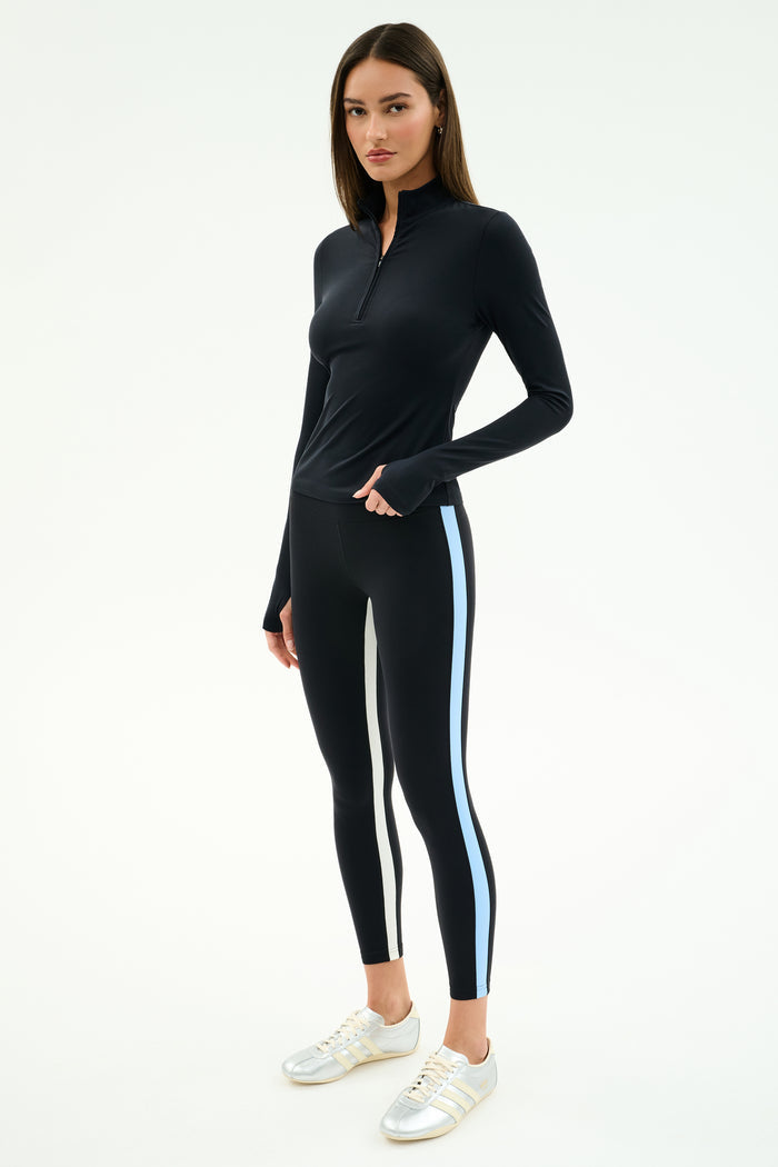 A woman wears the SPLITS59 Tess Rigor Half Zip Waist Length top in black with matching leggings featuring white and blue side stripes, and white sneakers, posed against a plain white background.