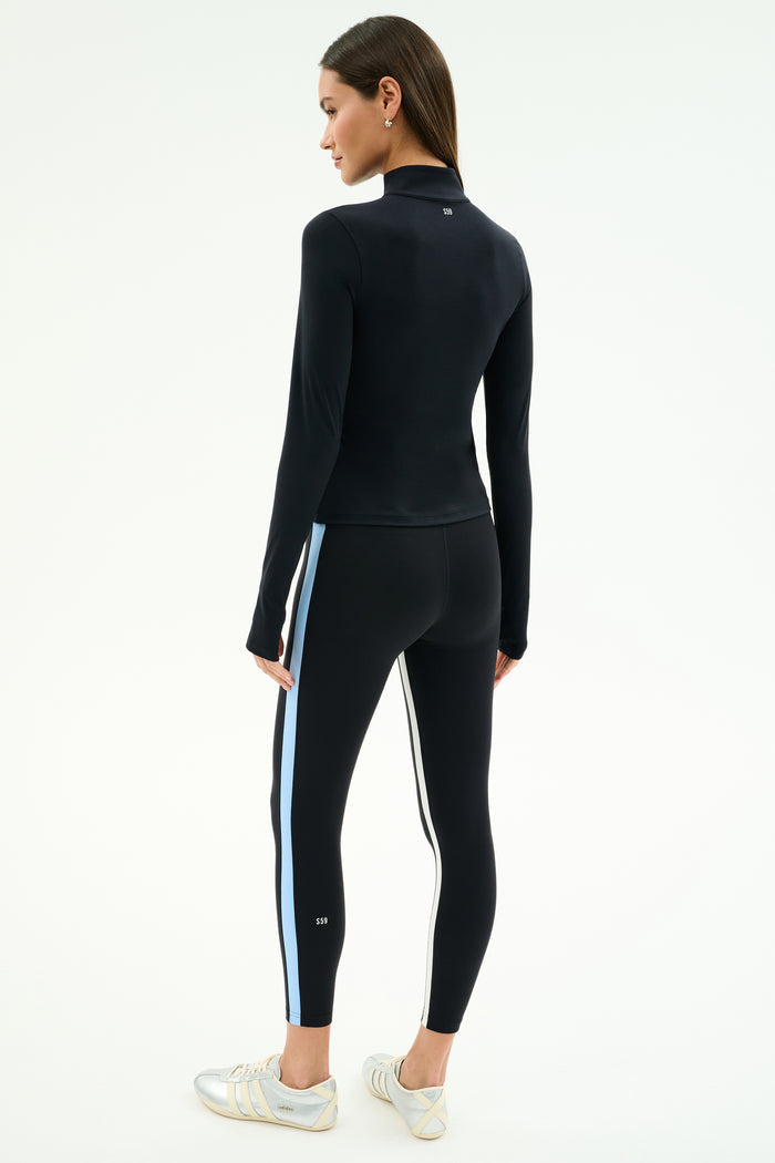 A woman faces away, wearing the SPLITS59 Tess Rigor Half Zip Waist Length top in black with matching leggings featuring white and blue side stripes, paired with white sneakers.
