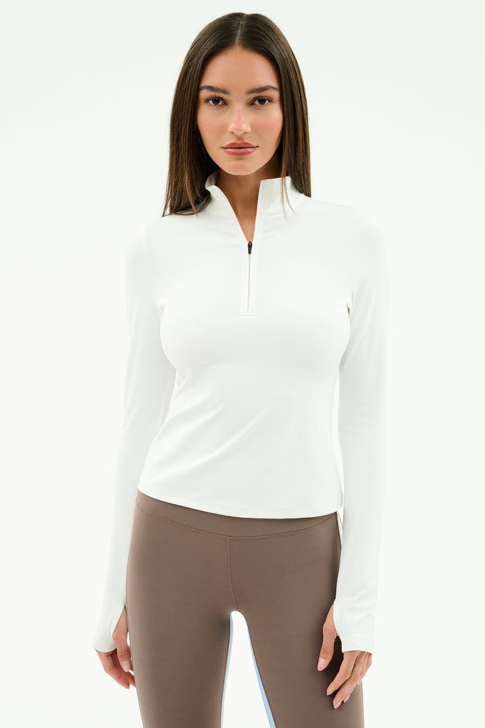 A woman faces forward against a plain white background, wearing the SPLITS59 Tess Rigor Half Zip Waist Length top in white and taupe nylon spandex leggings.