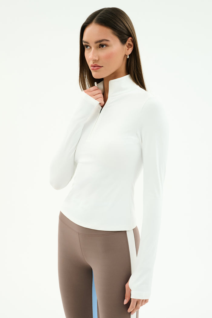 A woman stands against a white background wearing the SPLITS59 Tess Rigor Half Zip Waist Length top in white paired with taupe leggings.