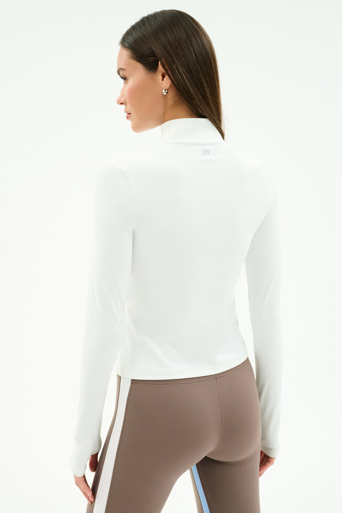 A woman faces away, wearing the SPLITS59 Tess Rigor Half Zip Waist Length top in white and high-waisted taupe leggings with white and blue side stripes made from stretchy nylon spandex.