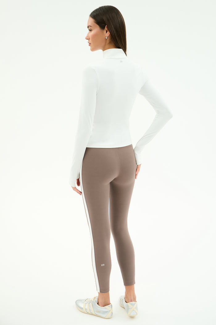 A woman stands facing away, wearing the SPLITS59 Tess Rigor Half Zip Waist Length top in white, taupe nylon spandex leggings with a white side stripe, and white sneakers against a plain background.