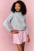 Young girl wearing a light blue hoodie with small patterns and a pink pleated skirt on a pink background