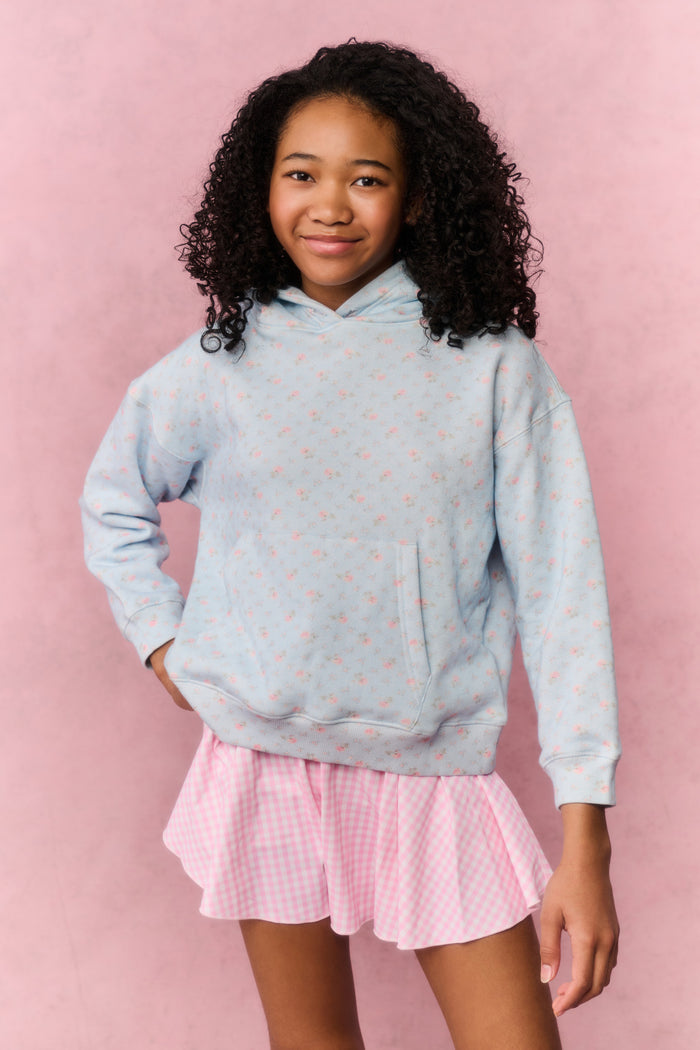 Young girl wearing a light blue hoodie with small patterns and a pink pleated skirt on a pink background