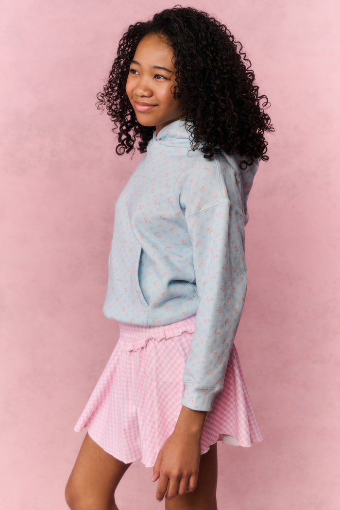 Young girl wearing a light blue hoodie with small patterns and a pink pleated skirt on a pink background