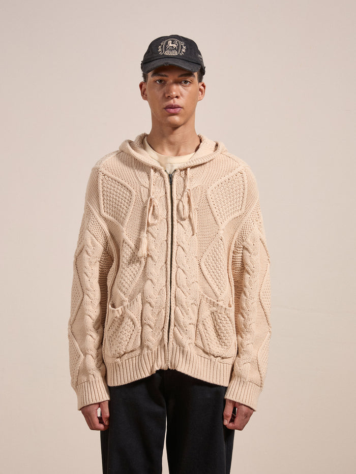 A person wears the Found Zip Up Cable Knit Hoodie in beige, a black cap with a white graphic, a beige shirt, and black pants while standing against a plain background. The cozy look and feel of the heavyweight cotton blend yarn of the hoodie adds to the overall comfortable vibe.